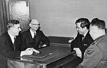 Four men sit facing each other around a table. Three of the men wear civilian suits, while one wears a military uniform