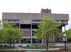 A topheavy concrete and brick building stands before a courtyard with paths and newly leafed trees.