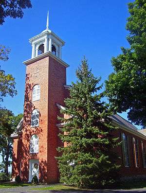 A brick building with a pine tree in front and a tower with white cupola