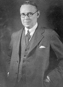 Studio portrait of Richard Gavin Reid. He has an oval, fleshy face with receding hair combed back, a short nose, small shaply mouth. His expression is quizzical and he is compressing a smile. He wears round horn-rimmed glasses and has his hands in the jacket pockets of his neat three-piece suit.