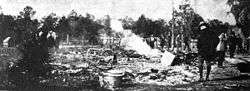 A black and white photograph of ashes from a burned building with several people standing nearby; trees in the distance