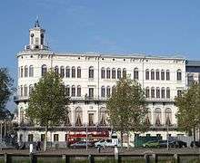 White building of four stories with a small tower on the top, green trees in the front