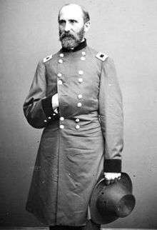 A balding white man with a full beard, wearing a long double-breasted military jacket. His right hand is in his jacket and his left is holding a wide-brimmed hat.