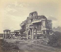 Ruins of a temple, entirely made of stone. The four-storied temple ruins rise behind two free-standing pillared structures, one of which hides the entrance to the temple. Sculptures of human forms are seen on the upper stories. Grass grows on various exposed surfaces of the ruins. A pathway, paved with stone slabs, fringes the visible perimeter of the temple.