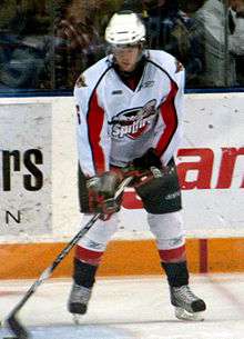 A teenage ice hockey player stickhandling a puck while standing still on the ice. His skates are shoulder-width apart and his eyes are downcast. He wears a white jersey with red and black trim, as well as a white, visored helmet.