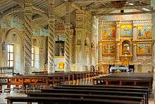 The nave of a church in three-quarter view and the left aisle with windows. The interior is dominated by white and bright orange colors. At the back, there is an altar with a statue surrounded by a five-paneled reredos.