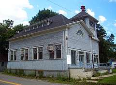 A blue building with a peaked roof, steps on the right and a large open tower at right rear with a conical red metal object on top.