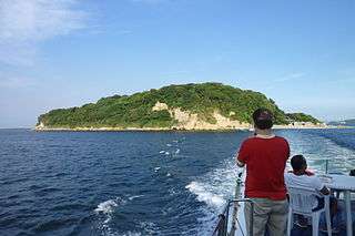 The island of Sarushima is golden on a late August day. In the lower right corner a few men on the deck of a boat look back at the island