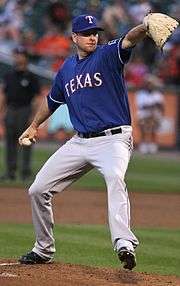 Pitcher in blue baseball cap coming out of his windup as he delivers a pitch to the plate