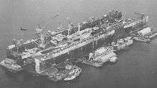 A floating drydock containing ships. It is surrounded by floating barges with workshops and a tugboat.