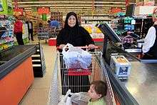 A woman stands behind a shopping cart at the checkout stand.