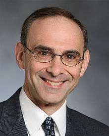 A Caucasian male with brown hair and eyeglasses, wearing a suit and tie, smiling at the camera.