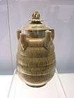 A tan jar with several rounded bands carved into the body and five upward facing tubes equadistantly spaced along the top area of the jar. The lid is crowned with a lotus flower carving.