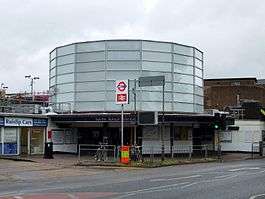 A white building with a rectangular, dark blue sign reading "SOUTH RUISLIP STATION" in white letters all under a light blue sky