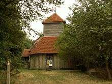 The end of a church seen between trees; the ground floor is timber framed, above this is a weatherboarded tower, and to the left part of the stone body of the church. All the roofs are covered in red tiles