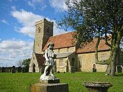 A stone church with red tiled roofs seen from an angle, the battlemented tower being on the left. In the foreground is a statue of a child.