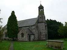 A small stone church seen from the northwest. On its right is a simple octagonal tower.  Immediately in front of the church is a yew tree and in the foreground to the right is a chest tomb