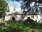 The base of a cross with four stone steps, in the shade of a yew tree, with the stone church in the background