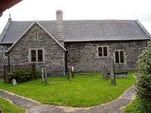 A very small, simple, single-story building with a gabled projection to the left. A few thin gravestones stand in front of the church