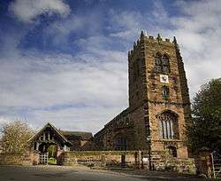 A sandstone church seen from the west with the tower prominent. In front of this is a wall leading to a lych gate on the left.  In front of the wall are stocks