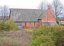 A small church in red brick with a grey roof seen from the side; protruding is a gabled wing with a chimney; the glass in the lancet windows is broken; bushes are in the foreground and leafless trees to the sides