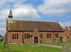 A small, simple, red sandstone church seen from the south. On its left is a small spirelet with a clock, and a small porch protrudes from the south wall