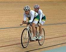 two women riding a tandem bicycle. The one in front, who is sighted, is acting as the pilot, while the second rider powers the bike.