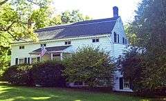 A white house with gray roof and large shrubs in front flying an American flag from the porch