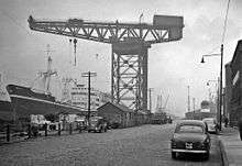 A black and white picture of a large crane reaching over a cargo ship.