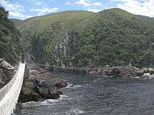 Suspension bridge at Storms River - River mouth between two ragged cliffs with some greenery, with the suspension bridge in the foreground