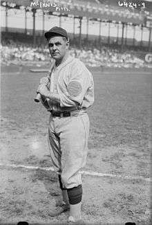 A black-and-white photo of a man wearing an old-style white baseball uniform and cap holding a baseball bat over his left shoulder