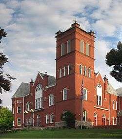 A two-story red brick building with a four-story square tower, stone foundation, and white-framed windows