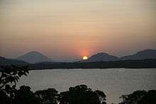 A partial view of a large lake, seen over a tree line. The further bank is covered by a thick forest, and the sun is setting over a mountain range in the background.