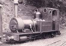 A small steam locomotive stands beside a banked area. On the left hand coal bunker is painted TAL-Y-LLYN RAILWAY. The track is covered with grass, and the rails are barely visible. In the foreground are some disused rails.