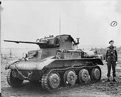 A small, squat tank sits in a field, its left-hand tracks facing the camera. Its gun barrel has been lengthened with a metal extension. A man in uniform with a black beret stands next to it. In the background, fields can be seen to the left, and several houses to the right.