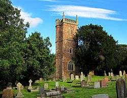 Stone building with arched windows and square tower, partially obscured by trees. Gravestones in the foreground