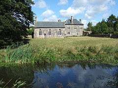 Seen beyond a river and a field is a simple stone chapel with three arched windows and a door; to the right is a two-storeyed stone house