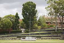 A grassy playing field with a war memorial at the end. Above the memorial are terraced steps leading up to trees and a chapel.