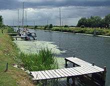 A broad, flat artificial cut with dark water and bright green raised banks.  A patch of green algae stains the water.  Several modern yachts are moored on wooden jetties spaced along the bank