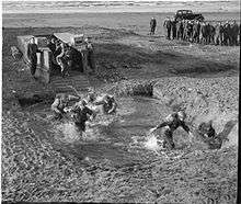 men on a beach wading through water are watched by a large group to their right