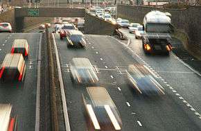 Photo of a busy two-lane dual carriageway from above, which abruptly drops away from view below a concrete underpass. An on-ramp is seen on the right and a traffic intersection is visible above the underpass.