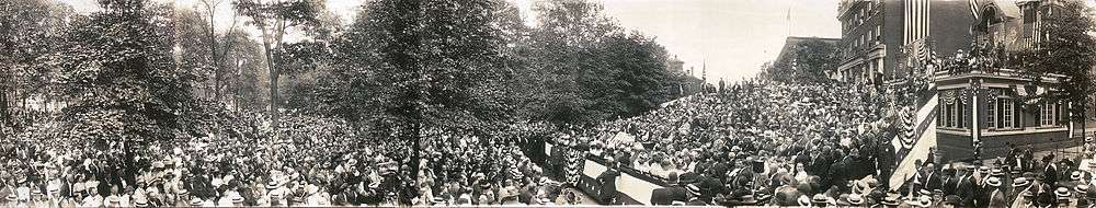 A panorama image showing a large crowd seated in chairs and standing outdoors around a speaker who delivers a speech on a podium in the center. Downtown brick buildings and large campaign posters are in the background.