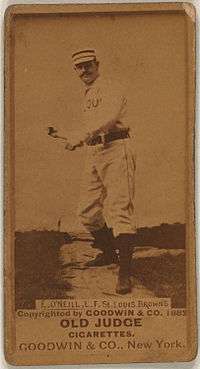 A man in a striped cap, white baseball uniform with the words "ST. LOUIS" obscured on the front and black belt pretends to swing an imaginary bat.