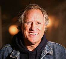 A Caucasian man wearing a denim jacket with blue eyes and dark blonde hair. Two orange lights are in the background.