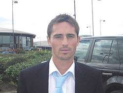 A head and shoulders of a man, wearing a black blazer, white shirt and blue tie.