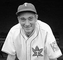 A man, wearing a baseball cap with a "T" in the center and a white baseball uniform with the Toronto Maple Leafs baseball logo on the left breast, leans forward smiling.