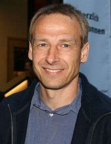 Head and shoulders of a man wearing an open-collar light blue shirt and a navy blue fleece. He has short blonde hair, blue eyes, and stubble.