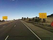 A highway near the top of a ridge. On either side of the highway are big yellow signs reading, "Trucks, Don't be Fooled—4 more miles of steep grades and sharp curves".