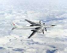 Top perpendicular starboard view of four-engine propeller-driven aircraft in-flight. The pale gray/white aircraft is flying above almost white terrain.