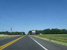 A two-lane road passing through farm fields.  A blue and white sign on the right indicates an ongoing construction project on US 113